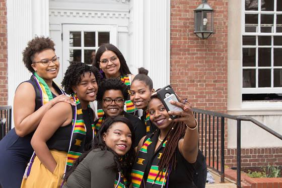 Photo of seven BIPOC Chatham University students in gramulticultural graduation stoles, posing for a selfie in front of James Laughlin Music Hall on Shadyside Campus